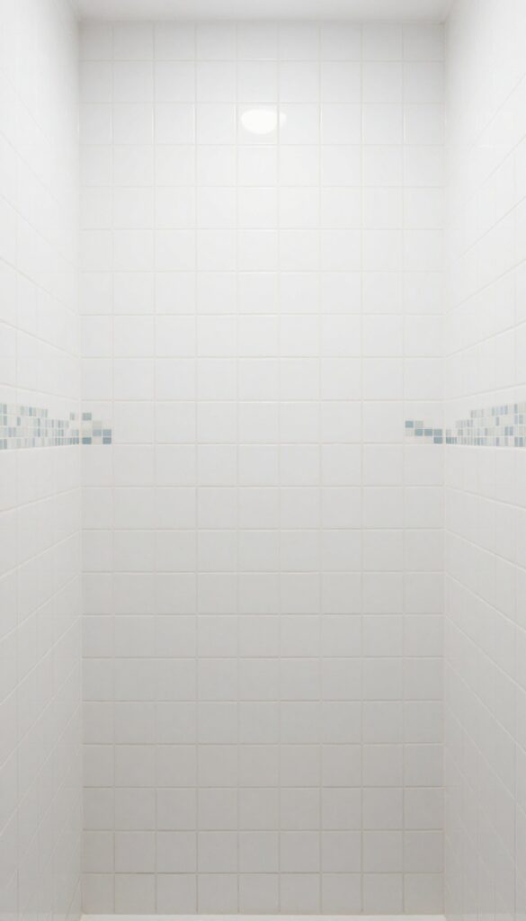 Close-up of a shower wall with plain white tiles featuring inlaid mosaic patterns in blue and gray, highlighting subtle texture and detail in natural light.