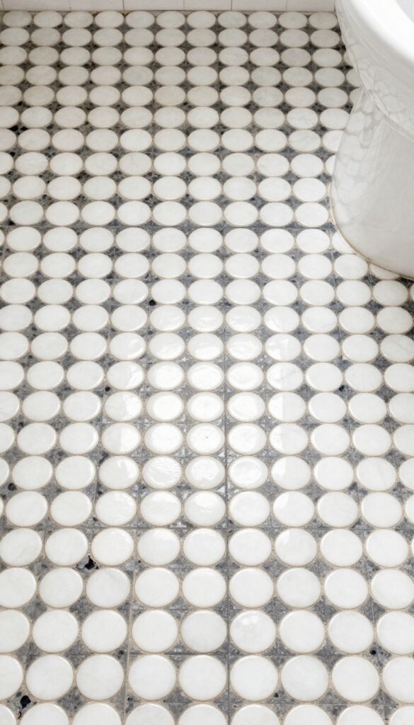 Close-up view of penny round tiles on a small bathroom floor, featuring white tiles with dark grout in natural light, highlighting the circular pattern and texture for a stylish, practical design.