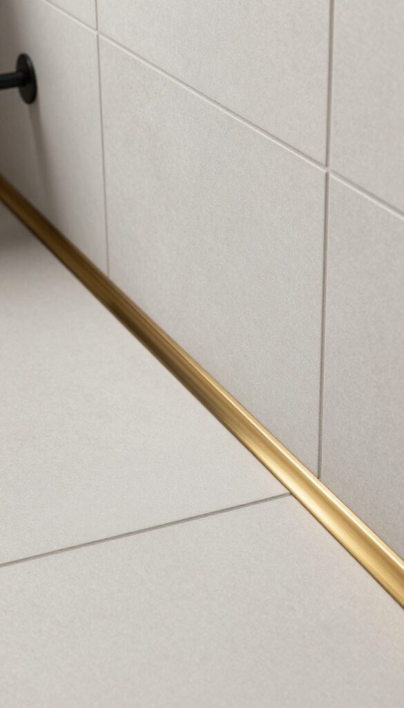 Close-up view of a shower threshold featuring thin brass metal strips for an industrial look, set in a modern bathroom with neutral tiles and natural lighting.