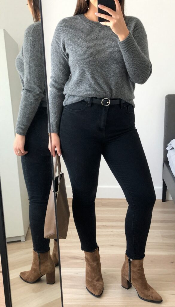 Woman in black skinny jeans and camel cashmere sweater with suede ankle boots, standing in a bright office or street setting.