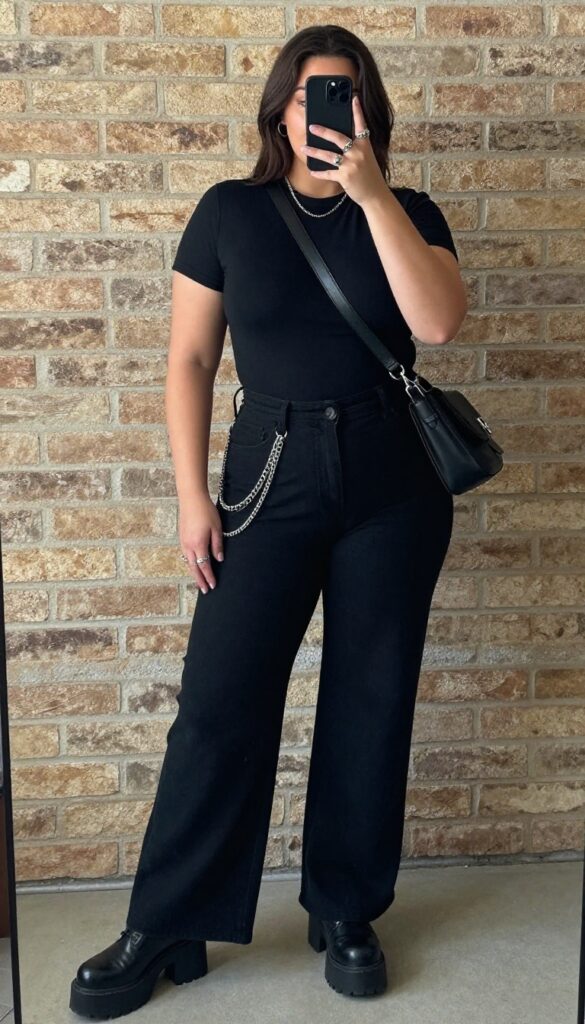 Plus-size woman in all-black grunge outfit with platform boots and layered necklaces, taking a mirror selfie.