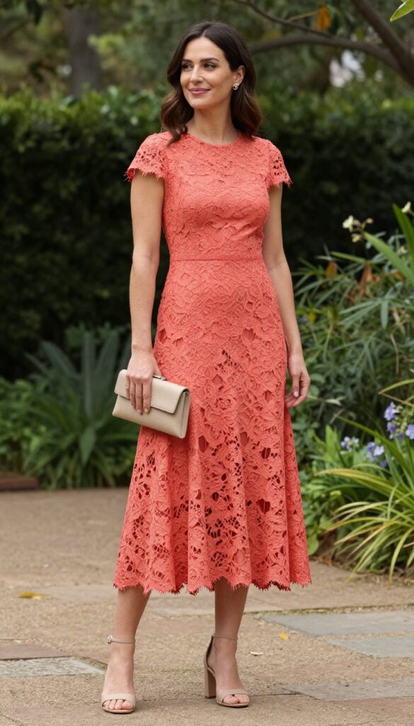 Woman in coral lace high-low hem dress with nude block heels and beige clutch at garden wedding