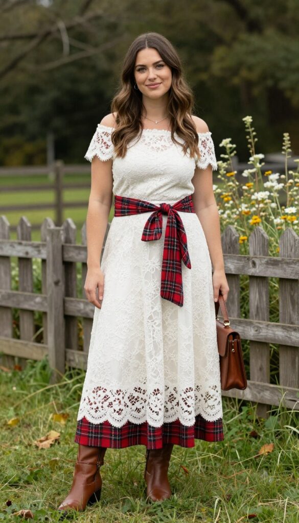Young adult woman in white lace off-shoulder dress with red plaid shirt tied at waist, brown ankle boots, and satchel, standing in grassy field with fence and wildflowers, smiling at camera.