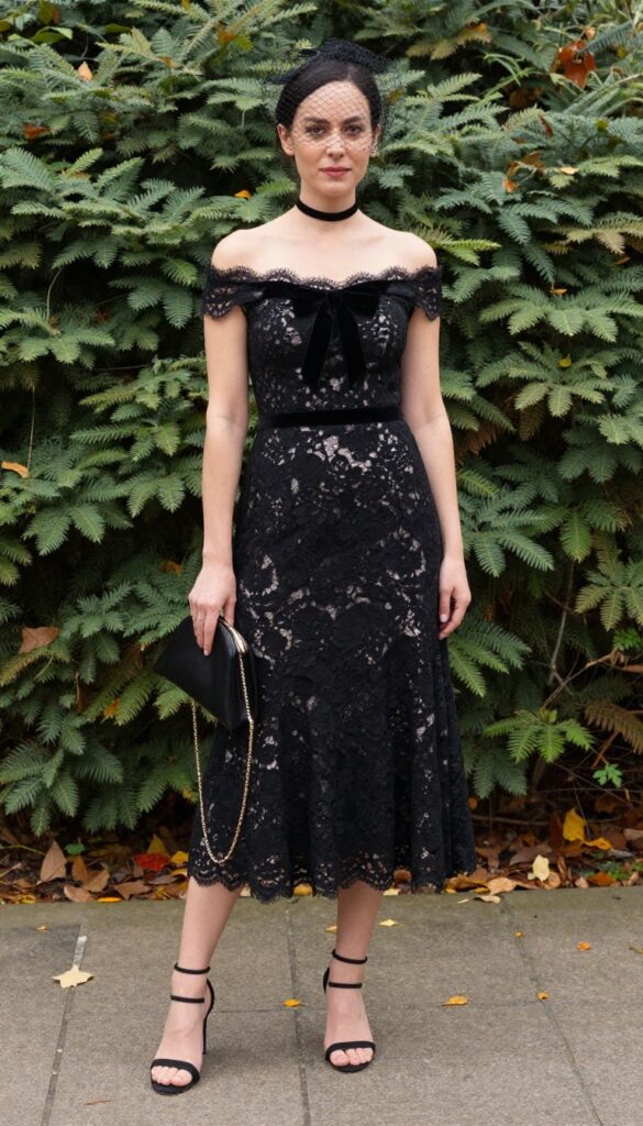 Young adult woman with pale skin and dark hair wearing off-shoulder black lace wedding dress with velvet choker bow, birdcage veil, and black strappy heels in autumn garden