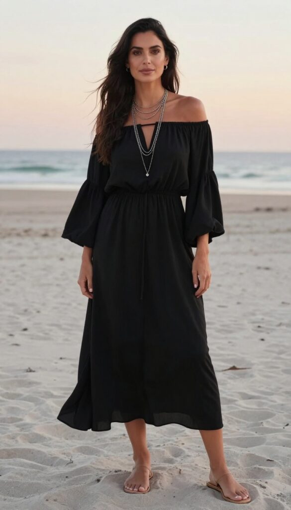 A young adult woman with fair skin and long dark hair wearing an off-shoulder black dress with wide bell sleeves, standing on a sandy beach at sunset, wearing layered silver necklaces and barefoot sandals, the dress flowing in the breeze.