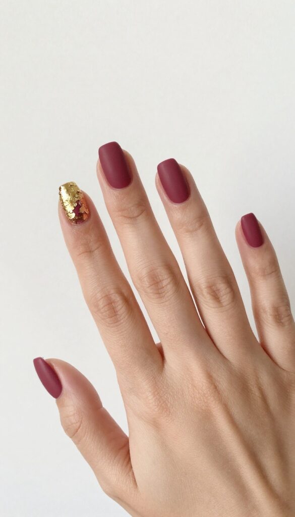 Matte cranberry nails with gold foil accents on ring finger