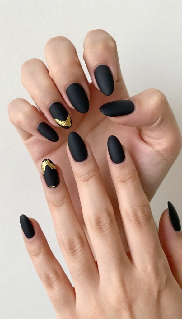 Almond nails with matte black polish and delicate gold foil accents near cuticles and tips