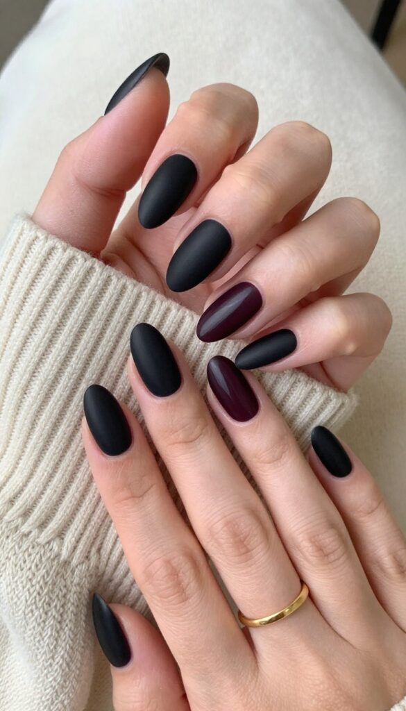 Matte black cherry nails with almond shape on a woman's hand, styled with a cream sweater and gold ring for a fall look.