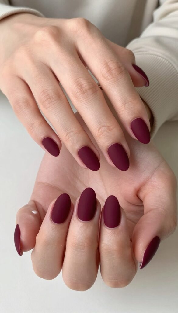 Velvet matte burgundy dip powder nails on almond shape, medium length, resting on cream sweater