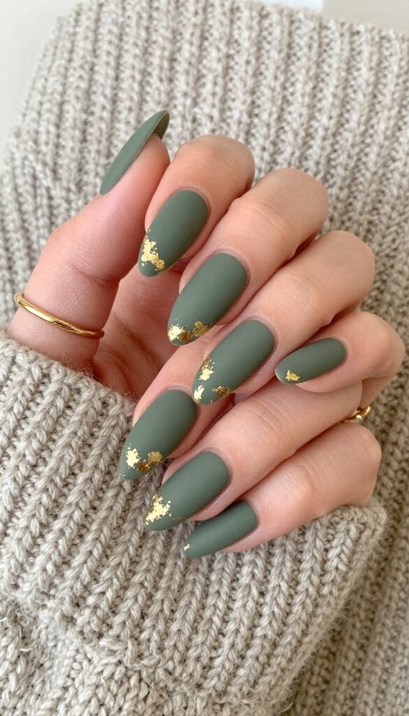 Matte olive green nails with gold foil accents on almond-shaped nails, styled with gold rings and a knit sweater for a chic fall look.